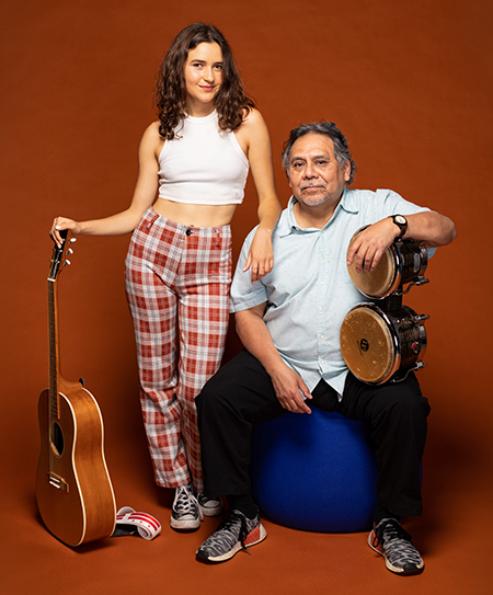 Anamaria Sayre, wearing red and white plaid pants and a white tank top, props up a guitar in one hand and uses the other hand to lean on the shoulder of her co-host Felix Contreras. Contreras, wearing a blue short sleeve button-up and black pants, is seated and holding a set of bongo drums. The pair is posed in front of a solid brown background.