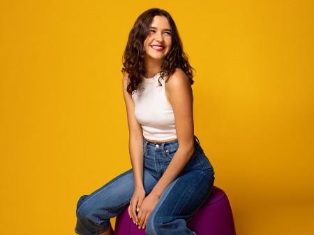 Anamaria Sayre wears a white tank top and jeans and sits on a magenta ottoman while looking away from the camera and smiling. The background of the image is a deep yellow-orange.