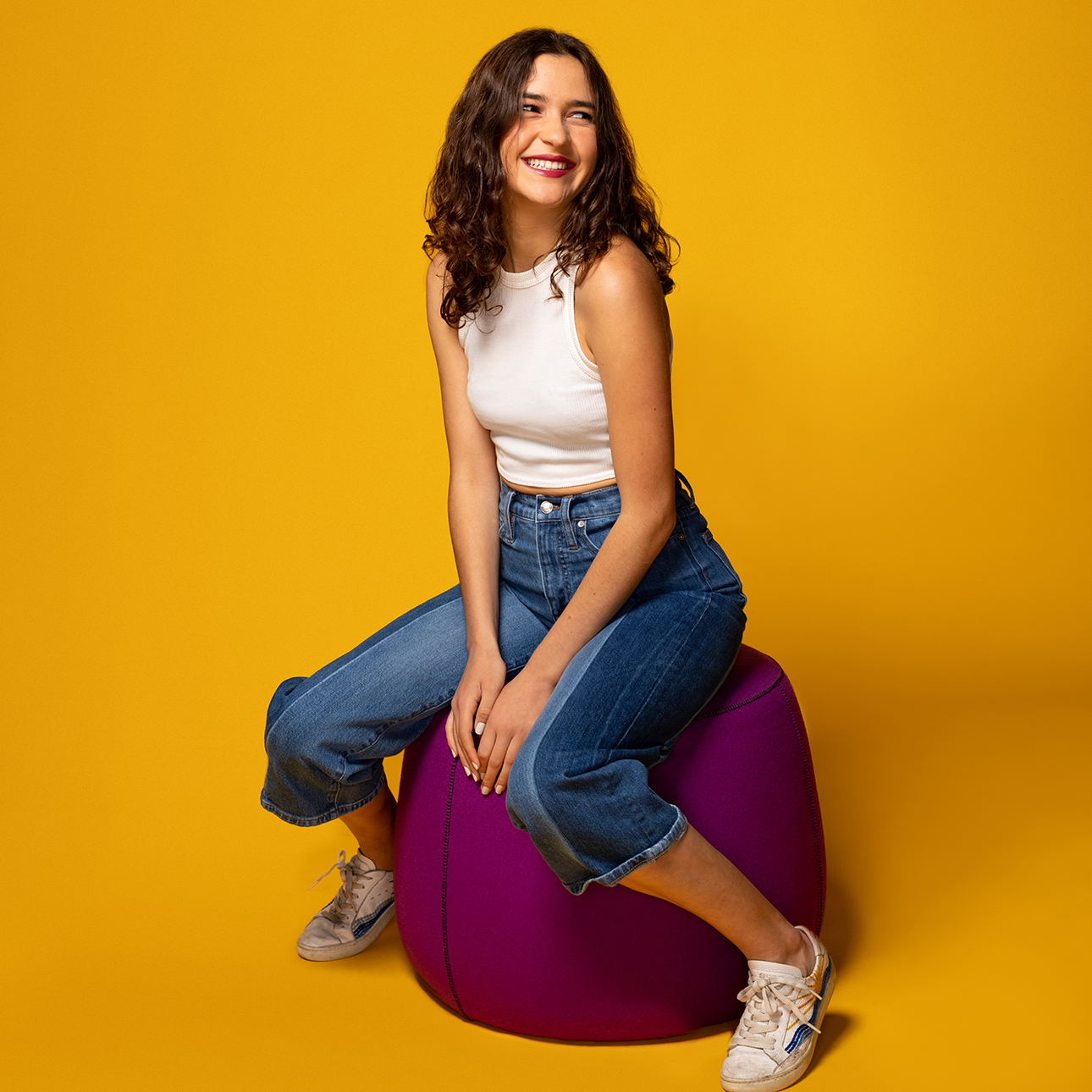 Anamaria Sayre wears a white tank top and jeans and sits on a magenta ottoman while looking away from the camera and smiling. The background of the image is a deep yellow-orange.