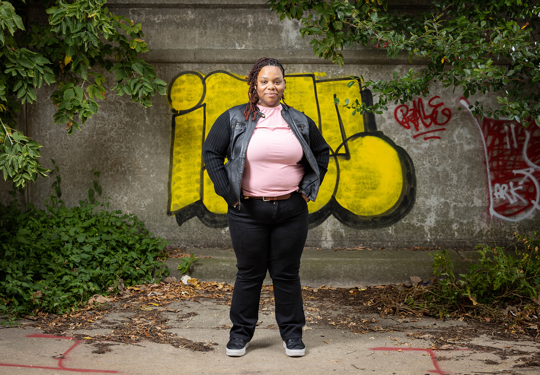  Alantha Miles stands outside in a pink top and black pants and jacket on a fall day. Behind her is a stone wall with graffiti and leaves on the ground.