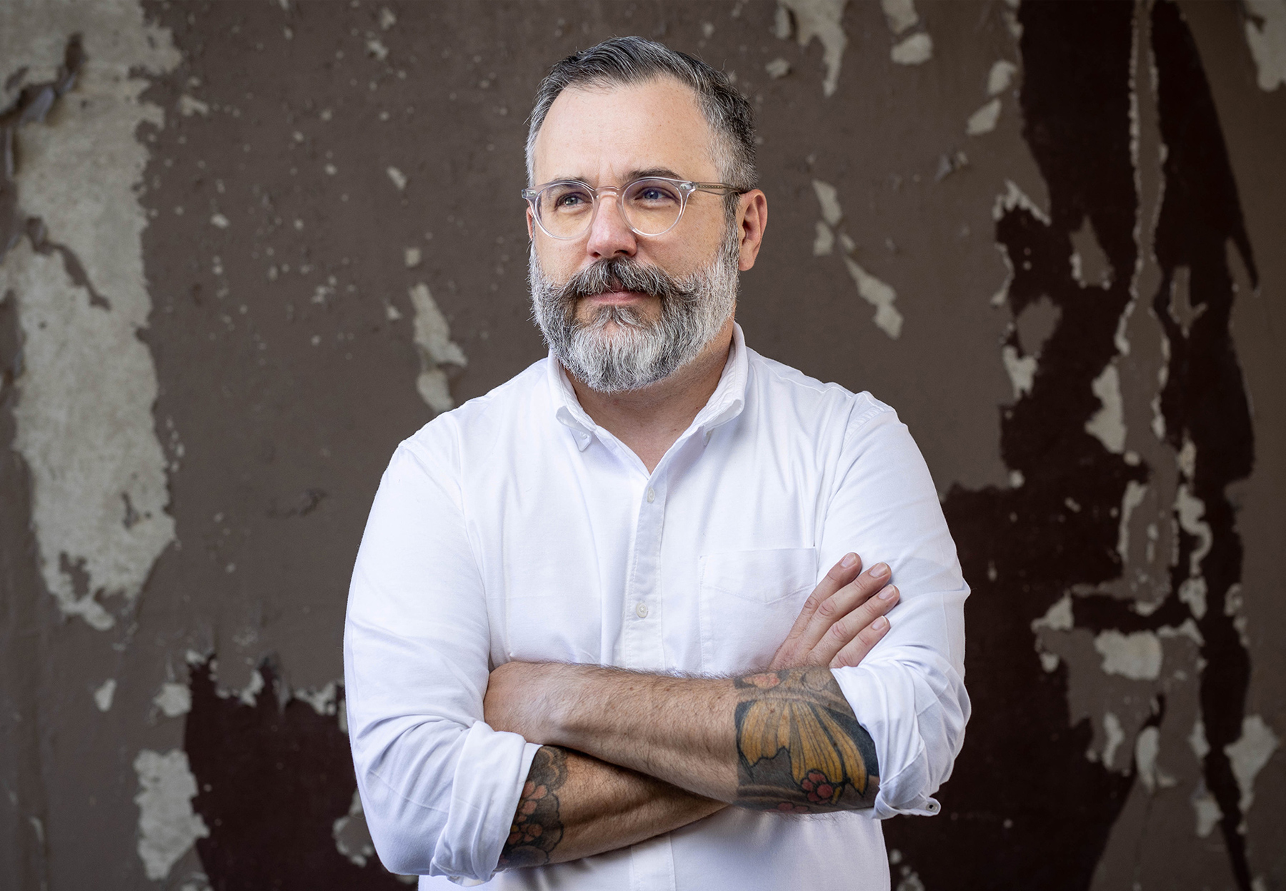 Andy Papachristos looks into the distance with his arms folded. He is wearing a white shirt and glasses and he has tattoos on his forearms. 