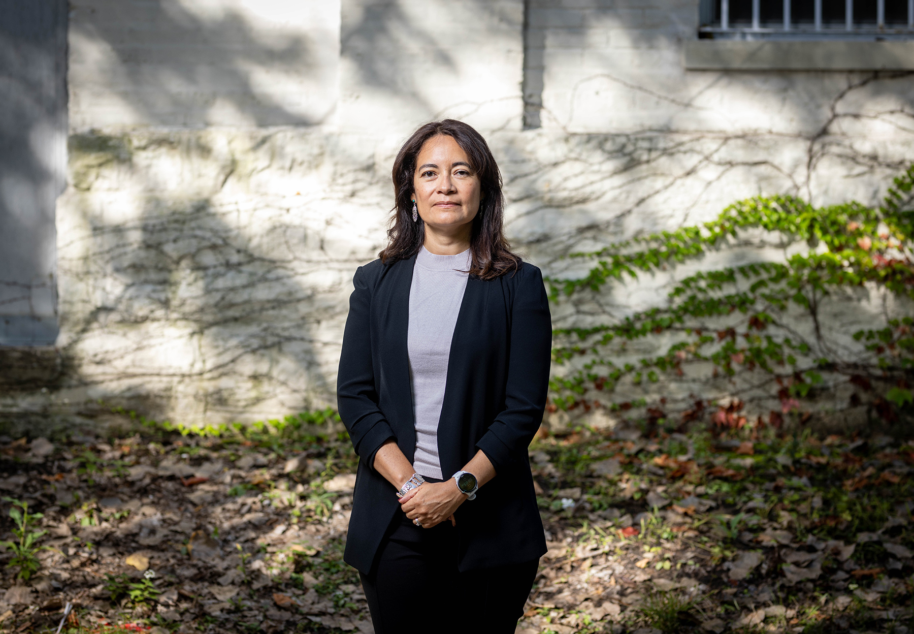 Soledad Adrianzén McGrath stands outside in a blue blazer and gray top on a fall day. She is looking directly at the camera and there is an ivy-covered brick building behind her.