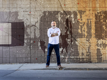 Professor Andrew Papachristos standing under a bridge in front of a wall with network science graphics surrounding him. He is wearing a white shirt and dark jeans and has his arms crossed over his chest.