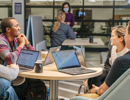 Four students sit at a round table with laptops in front of them, chatting with each other.