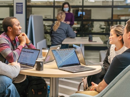 Four students sit at a round table with laptops in front of them, chatting with each other.