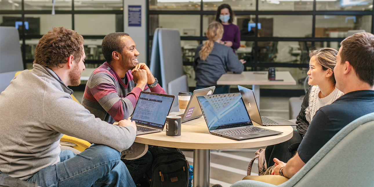 Four students sit at a round table with laptops in front of them, chatting with each other.