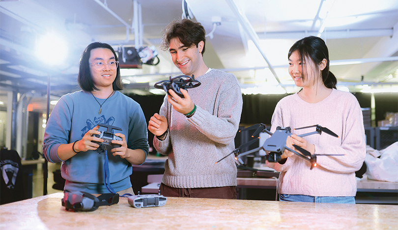 Three students stand at a workbench holding drones. 