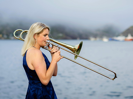 Hillary Simms, wearing a blue dress, plays the trombone in front of a foggy lake.