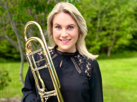 Hillary Simms, wearing a black blouse and jeans, smiles at the camera while holding a trombone in a grassy landscape.