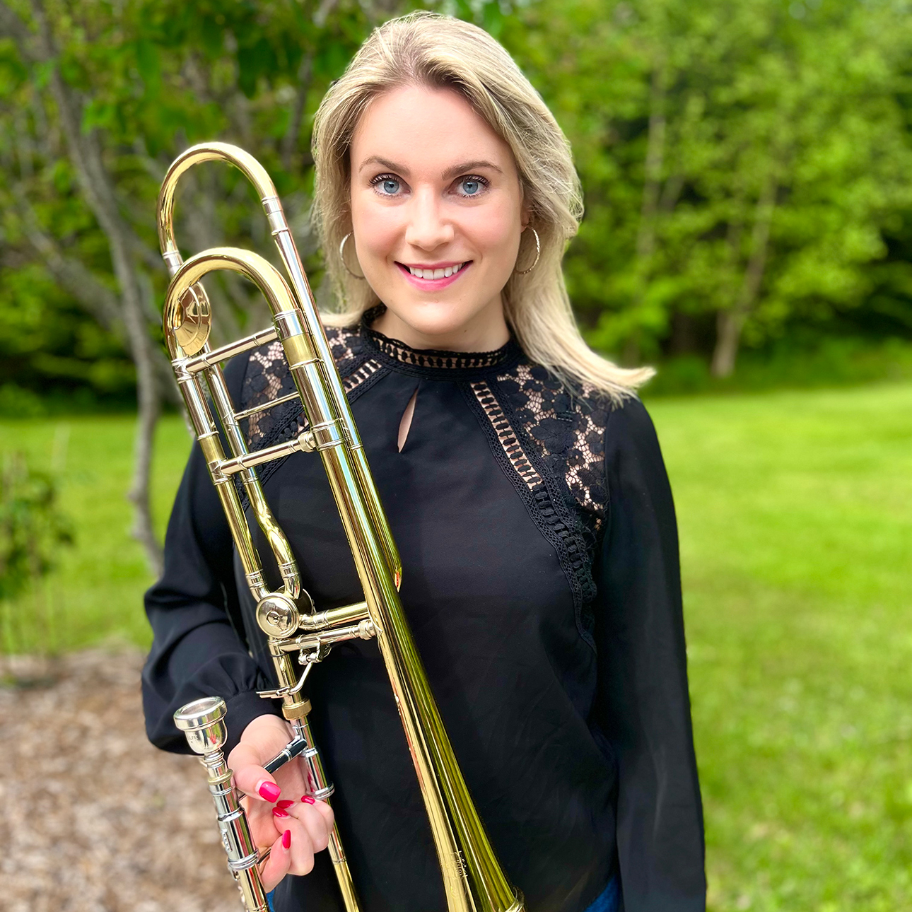 Hillary Simms, wearing a black blouse and jeans, smiles at the camera while holding a trombone in a grassy landscape.