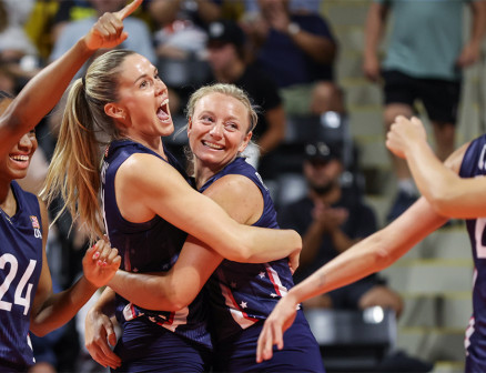 LOVB Pro players Annie Drews and Jordyn Poulter hug in celebration on the court, while teammate Chiaka Ogbogu points and smiles at their teammates in the foreground.