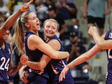 LOVB Pro players Annie Drews and Jordyn Poulter hug in celebration on the court, while teammate Chiaka Ogbogu points and smiles at their teammates in the foreground.