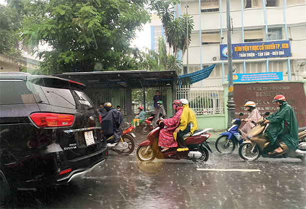 Several people are wearing rain jackets and riding motorbikes on a busy street in Ho Chi Minh City, Vietnam, while it is pouring rain.