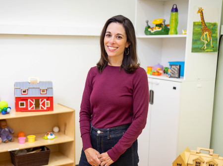 Megan York Roberts, wearing a purple long-sleeve top and jeans, stands smiling in a classroom full of children’s toys.