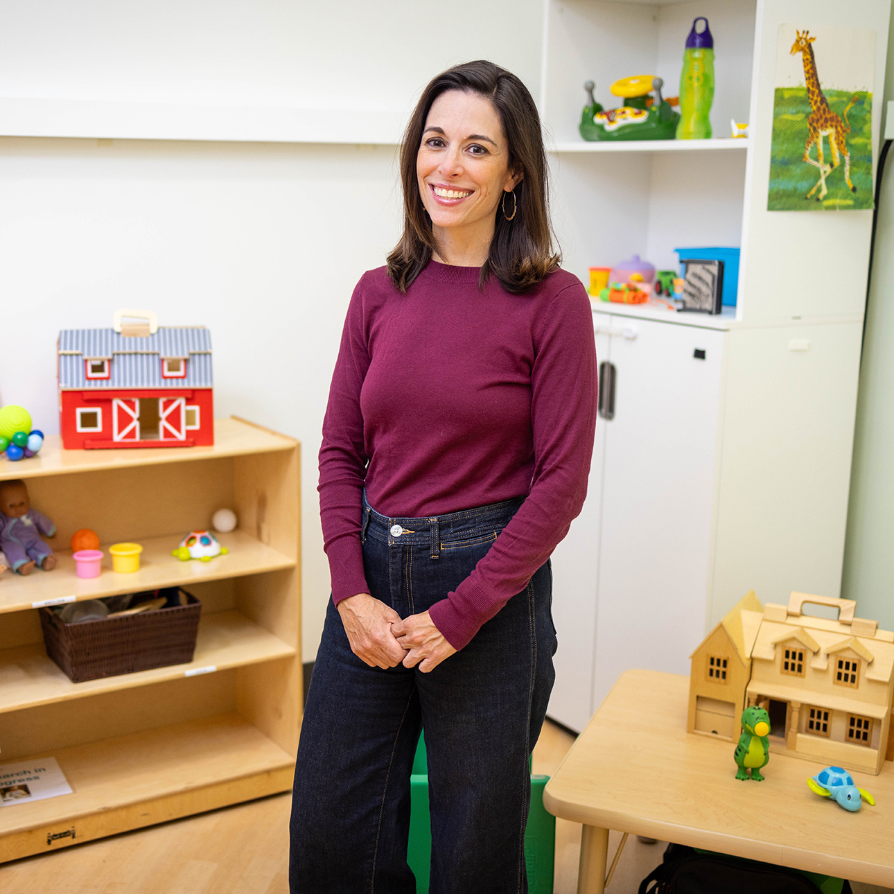 Megan York Roberts, wearing a purple long-sleeve top and jeans, stands smiling in a classroom full of children’s toys.