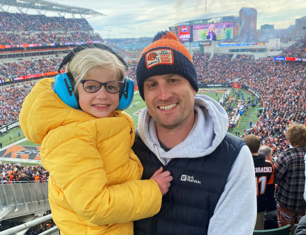 Mike Schroder is in the stands of a Bengal Tigers football game. He is wearing a Bengal Tigers hat, sweatshirt and vest and holds his daughter Ella in his arms. Ella wears a bright yellow jacket.