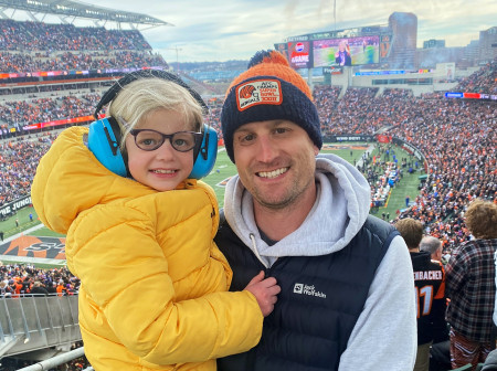 Mike Schroder is in the stands of a Bengal Tigers football game. He is wearing a Bengal Tigers hat, sweatshirt and vest and holds his daughter Ella in his arms. Ella wears a bright yellow jacket.