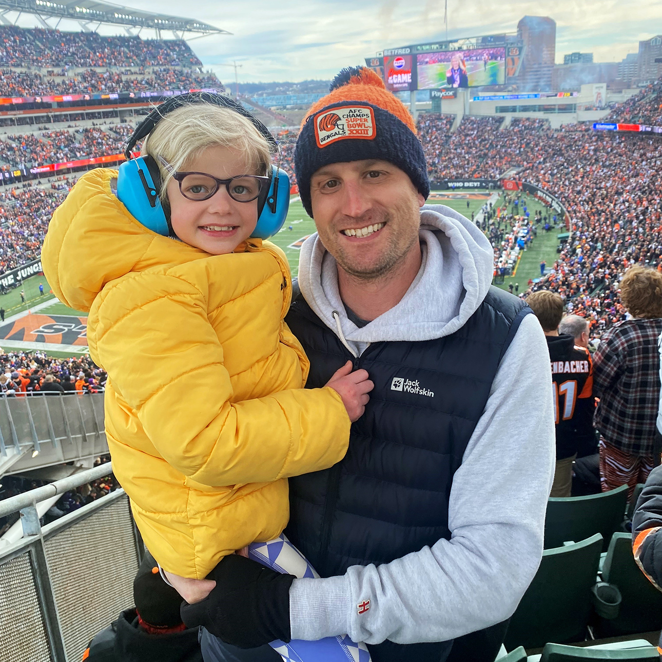 Mike Schroder is in the stands of a Bengal Tigers football game. He is wearing a Bengal Tigers hat, sweatshirt and vest and holds his daughter Ella in his arms. Ella wears a bright yellow jacket.