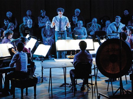 Alan Pierson stands on stage in front of a pianist and other musicians with his arms poised to conduct while an audience looks on behind him.