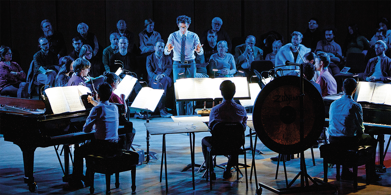 Alan Pierson stands on stage in front of a pianist and other musicians with his arms poised to conduct while an audience looks on behind him.