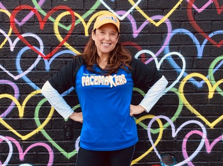 Susan Avery, wearing her blue and black Pacemakers uniform and a yellow hat, stands smiling with her hands on her hips in front of a black mural with colorful hearts.