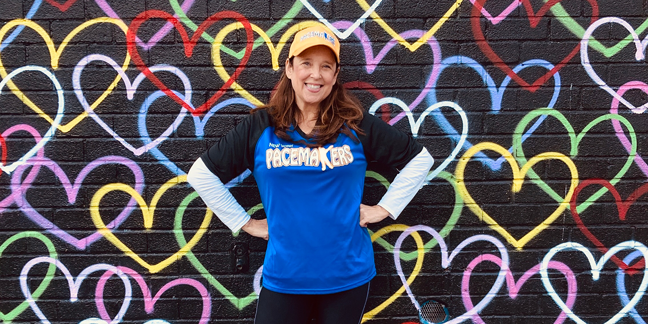 Susan Avery, wearing her blue and black Pacemakers uniform and a yellow hat, stands smiling with her hands on her hips in front of a black mural with colorful hearts.