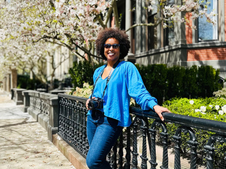 Jehana Ray leans against a fence on a residential street, wearing a blue blouse and jeans with a camera hanging around her neck.