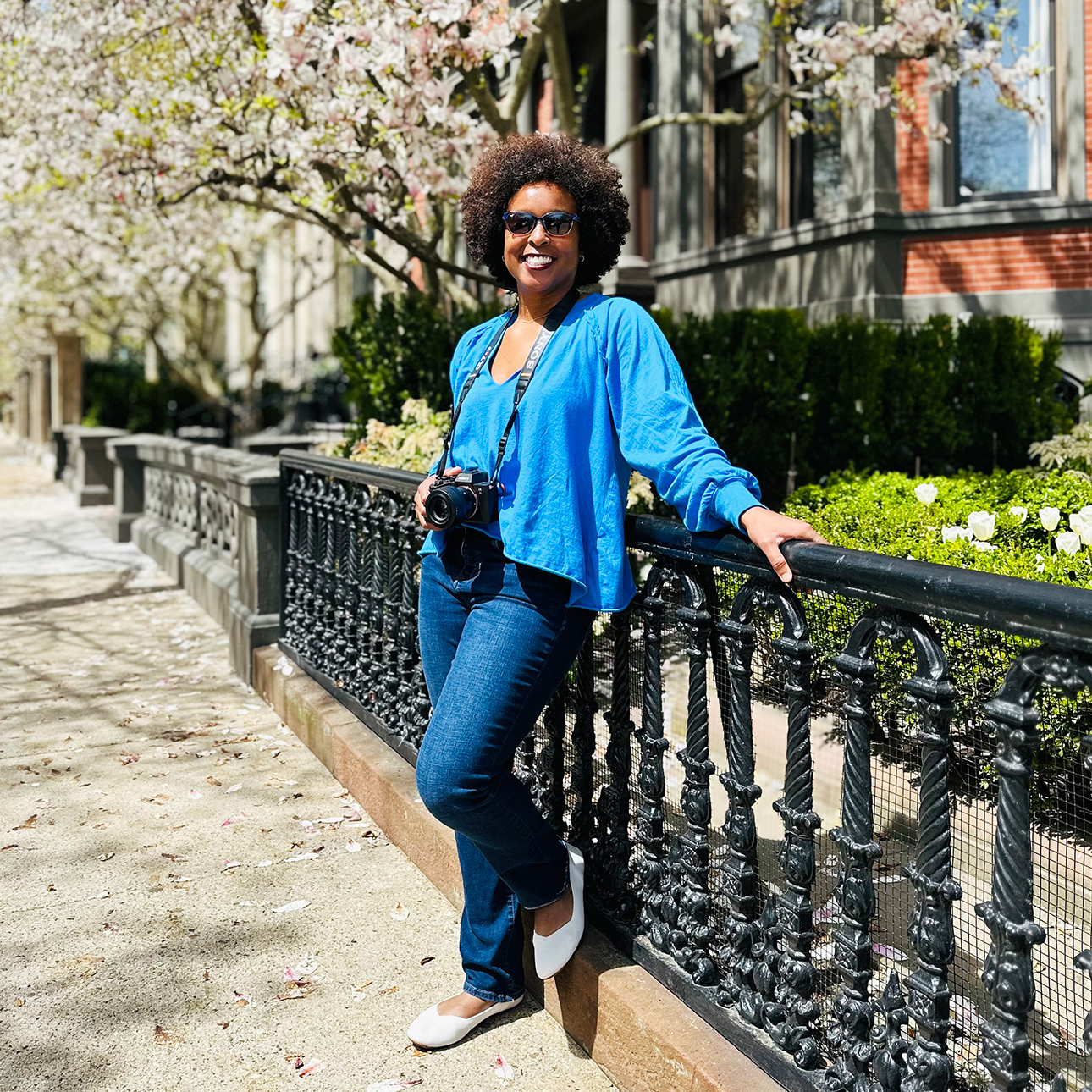 Jehana Ray leans against a fence on a residential street, wearing a blue blouse and jeans with a camera hanging around her neck.