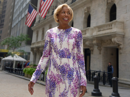 Sharon Bowen wears a white dress patterned with purple and pink flower blossoms cascading down it. She stands outside the New York Stock Exchange and smiles as she looks away from the camera. American flags can be seen on the building behind her.