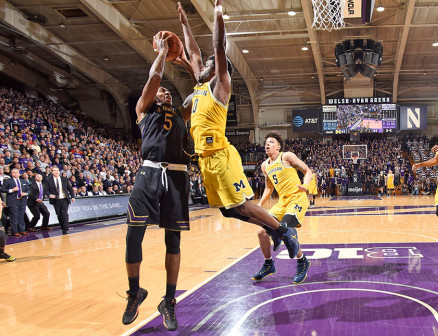 Dererk Pardon, in a black Northwestern basketball jersey, shoots a lay-up in a packed Welsh-Ryan Arena as a player from the University of Michigan, dressed in a yellow jersey, jumps to block him. One Northwestern player and two Michigan players approach in the background.