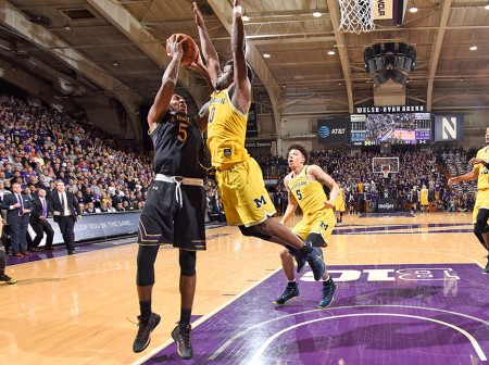 Dererk Pardon, in a black Northwestern basketball jersey, shoots a lay-up in a packed Welsh-Ryan Arena as a player from the University of Michigan, dressed in a yellow jersey, jumps to block him. One Northwestern player and two Michigan players approach in the background.