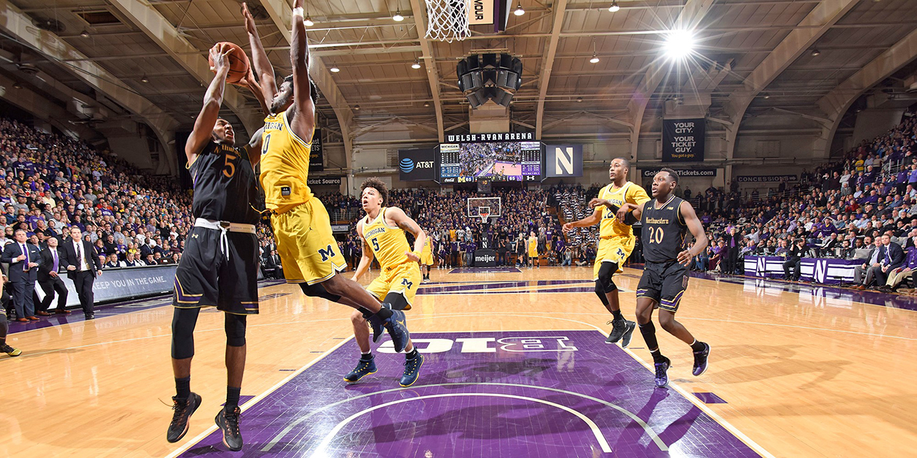 Dererk Pardon, in a black Northwestern basketball jersey, shoots a lay-up in a packed Welsh-Ryan Arena as a player from the University of Michigan, dressed in a yellow jersey, jumps to block him. One Northwestern player and two Michigan players approach in the background.