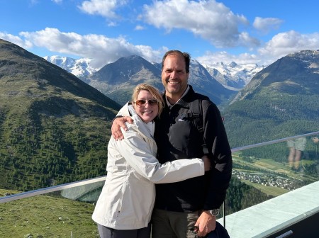 Kate Polozie, who is wearing a white coat, dark pants and sunglasses, hugs Stephen Polozie, who wears a black jacket and green shorts. The pair are posing on a balcony that overlooks a green, mountainous landscape.