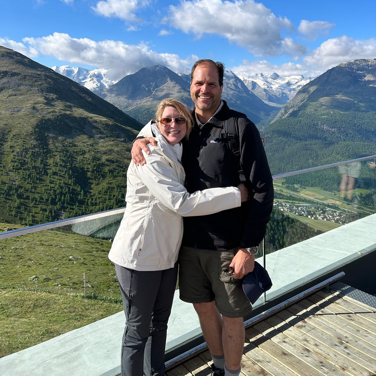 Kate Polozie, who is wearing a white coat, dark pants and sunglasses, hugs Stephen Polozie, who wears a black jacket and green shorts. The pair are posing on a balcony that overlooks a green, mountainous landscape.