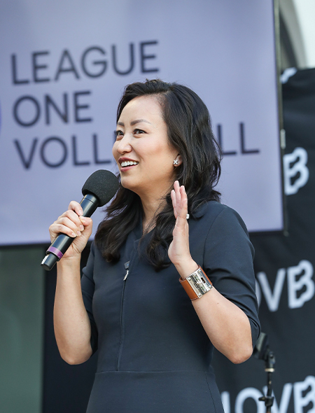 Gao, wearing a black dress, holds a microphone and gestures with her left hand. Gao is standing in front of a backdrop that reads “League One Volleyball.”