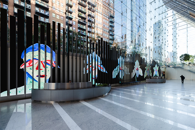 A wide shot of the outdoor video installation Overheard in the Underworld against a backdrop of Chicago buildings. Tall, vertical video panels are lined up in a row and each one shows a different piece of a horizontal image that shows a humanoid head morphing into a flower.