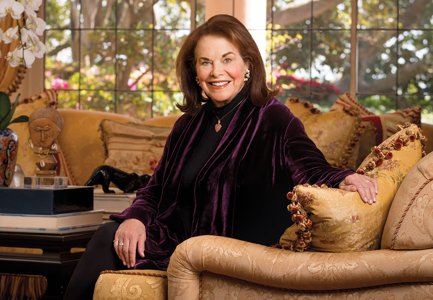 Sherry Lansing smiles, sitting on a couch with one arm rested on a pillow.