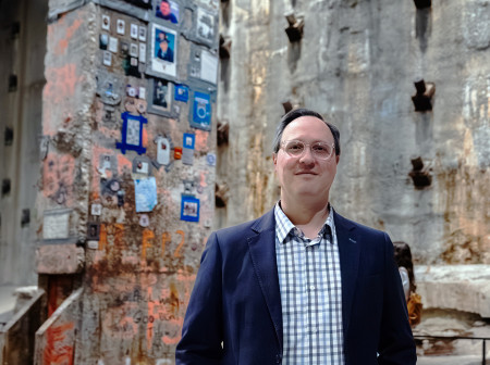 Ian Kerrigan stands in front of a cement wall peppered with small images and graffiti. 