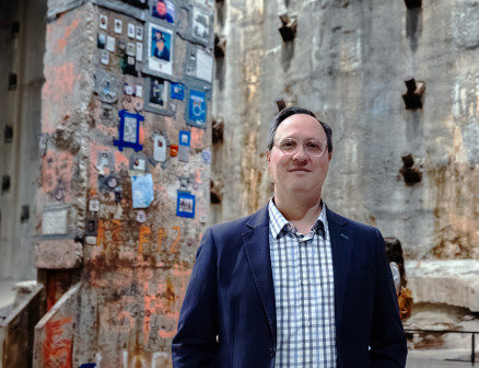 Ian Kerrigan stands in front of a cement wall peppered with small images and graffiti. 