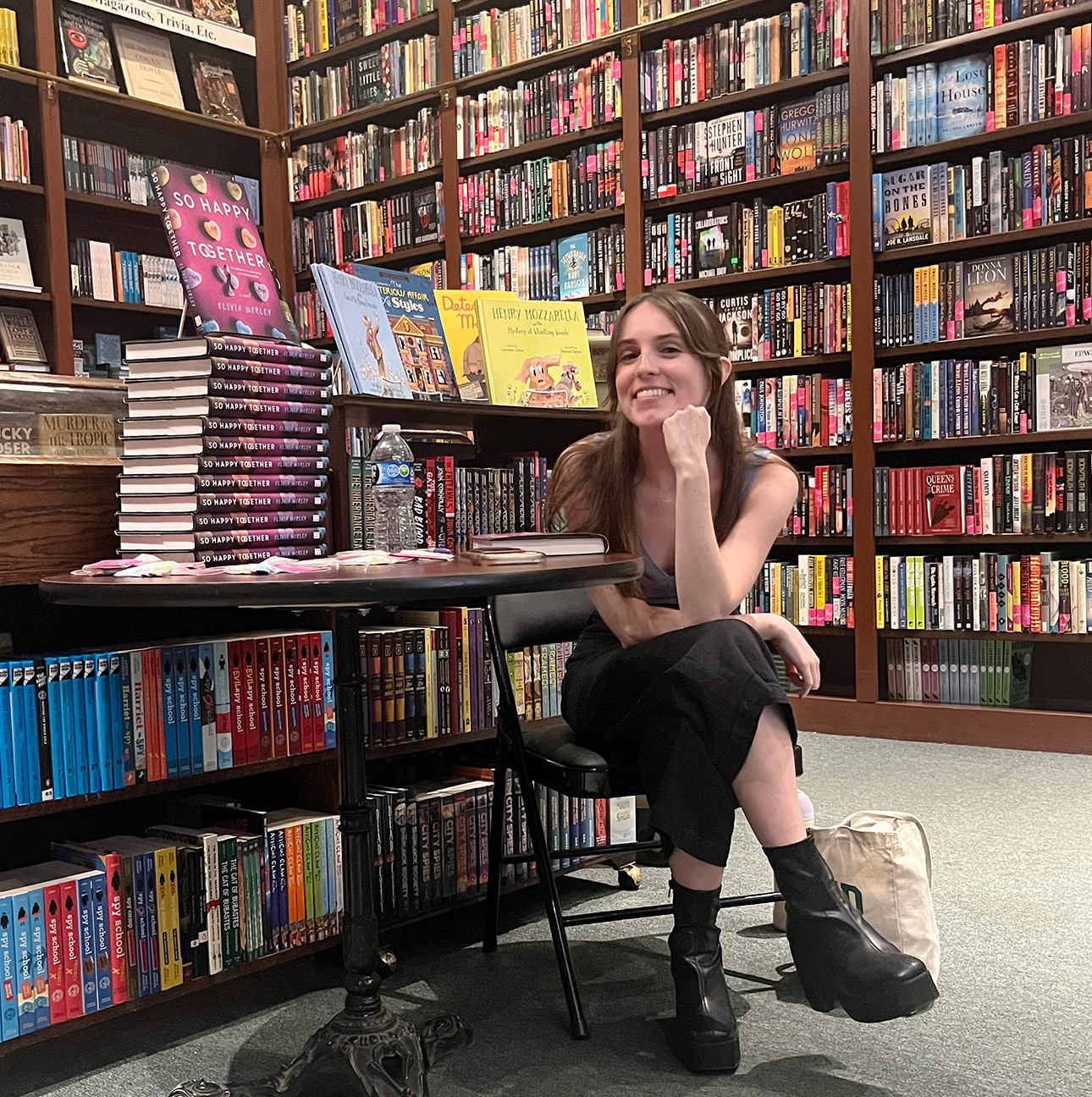 Surrounded by books in a bookstore, a woman sits on a chair with her legs crossed, leaning over with her elbow on her leg and fist under her chin.
