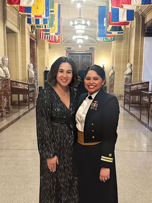 Two women, one of whom is wearing military dress, pose together, smiling in a hallway with flags hanging from the ceiling.