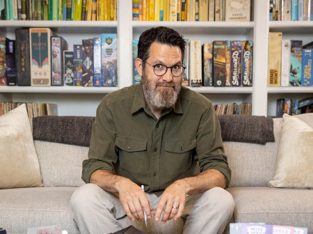 Sandy Weisz looks at the camera while sitting on a couch in front of some bookshelves. In front of him is a coffee table laden with puzzle-hunt paraphernalia.