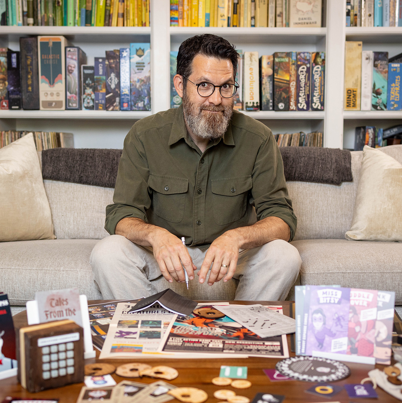 Sandy Weisz looks at the camera while sitting on a couch in front of some bookshelves. In front of him is a coffee table laden with puzzle-hunt paraphernalia.