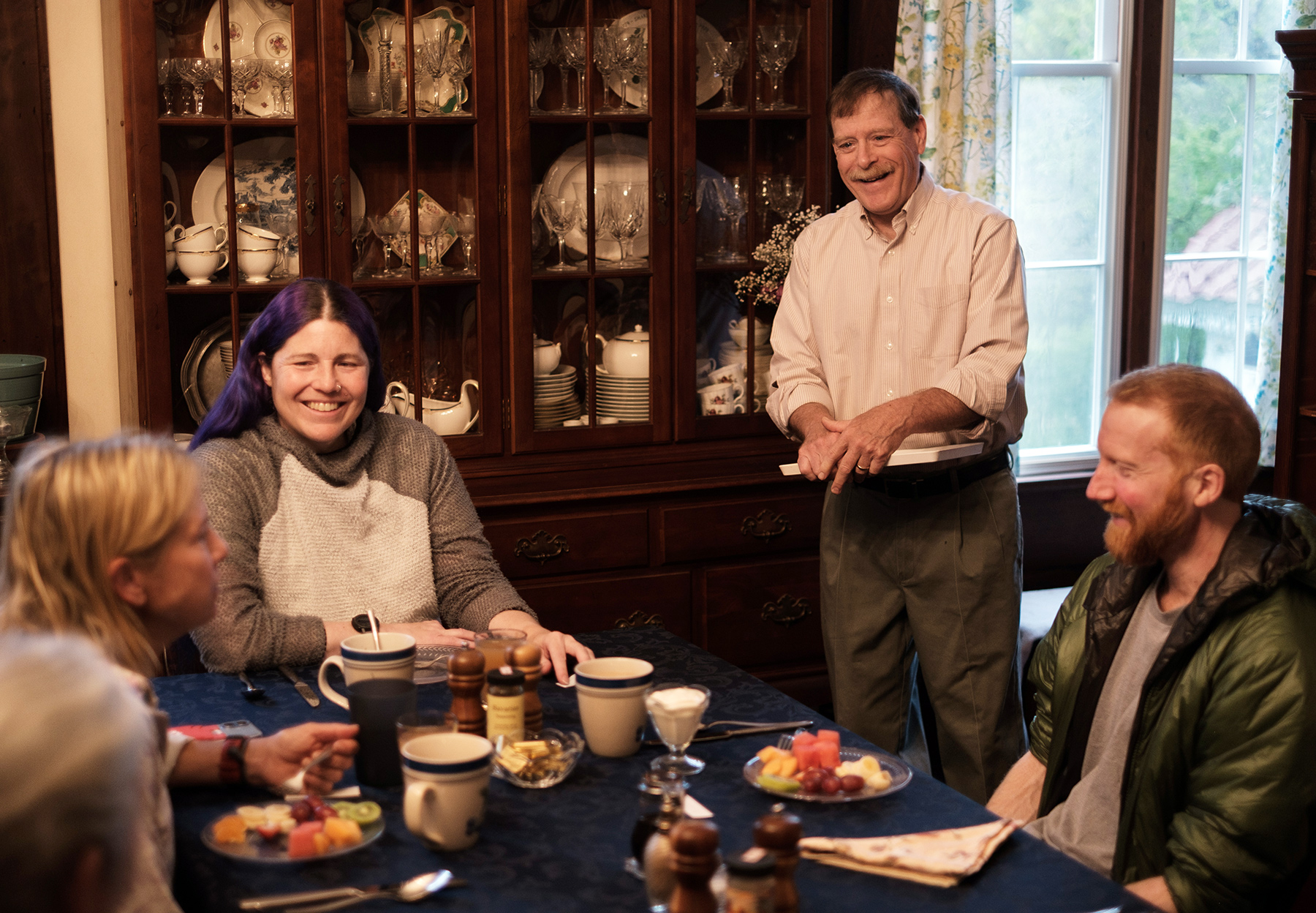 A group of guests sit around a table eating breakfast while Scott Jenkins stands nearby chatting with them.