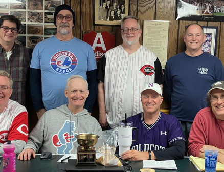 A group of men wearing baseball jerseys stand and sit in two rows at a table.