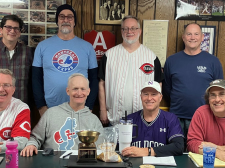 A group of men wearing baseball jerseys stand and sit in two rows at a table.