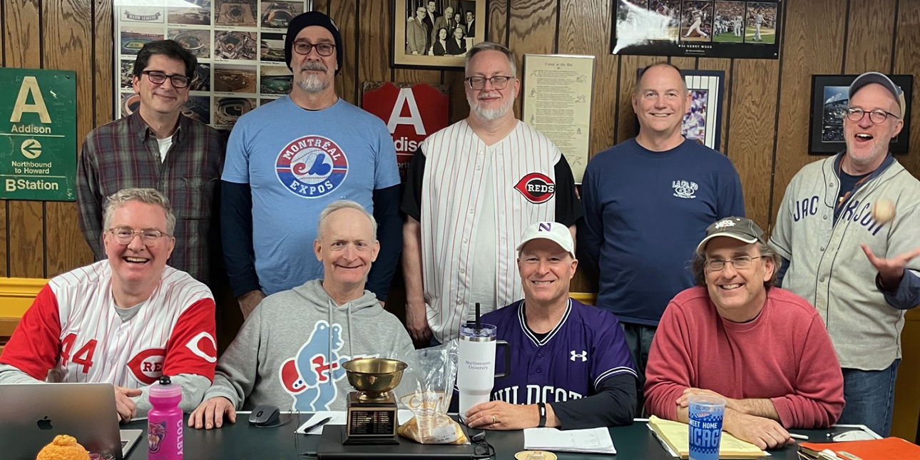 A group of men wearing baseball jerseys stand and sit in two rows at a table.
