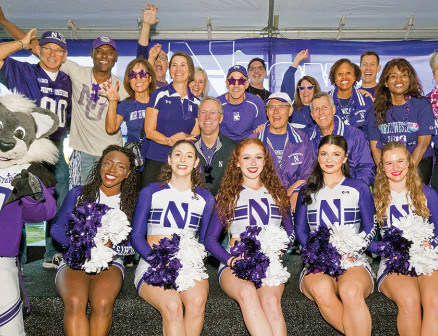 Six members of the Northwestern Spirit Squad, dressed in uniform and holding pom poms, sit on a bench next to Willie the Wildcat and in front of a group of smiling alumni who are raising their hands in celebration and to form wildcat paws.