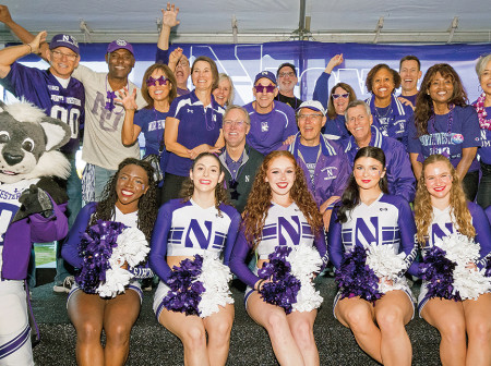 Six members of the Northwestern Spirit Squad, dressed in uniform and holding pom poms, sit on a bench next to Willie the Wildcat and in front of a group of smiling alumni who are raising their hands in celebration and to form wildcat paws.