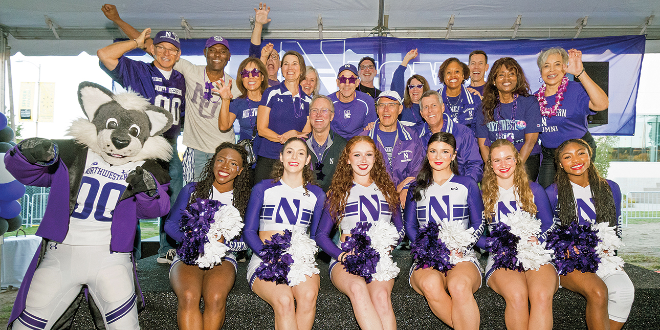 Six members of the Northwestern Spirit Squad, dressed in uniform and holding pom poms, sit on a bench next to Willie the Wildcat and in front of a group of smiling alumni who are raising their hands in celebration and to form wildcat paws.
